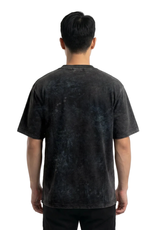 A man viewed from behind wearing a dark charcoal gray mineral-wash t-shirt and black pants, standing against a plain white studio background.