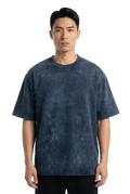 A man wearing a dark blue acid-wash t-shirt and black pants, standing against a plain white studio background.
