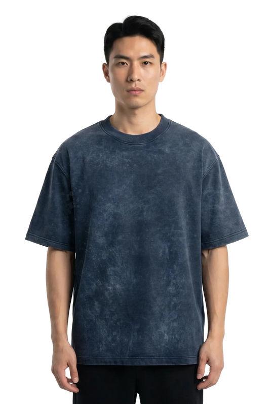 A man wearing a dark blue acid-wash t-shirt and black pants, standing against a plain white studio background.